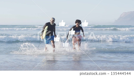 Running senior man and woman carrying yellow surfboard from surf at shoreline, wearing rash guards 135444544