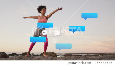 Balancing woman wearing coral sports bra doing yoga on rocky shore at sunset, blue speech bubbles 135444576