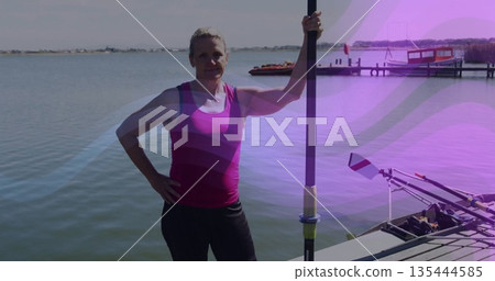 Standing woman wearing pink athletic top and black leggings on dock, holding rowing oar with shell Standing woman wearing pink athletic top and black leggings on dock, holding rowing oar with shell 135444585