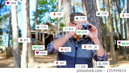 Photographer in quilted vest taking photos in pine forest clearing, with DSLR notification icons Photographer in quilted vest taking photos in pine forest clearing, with DSLR notification icons 135444614