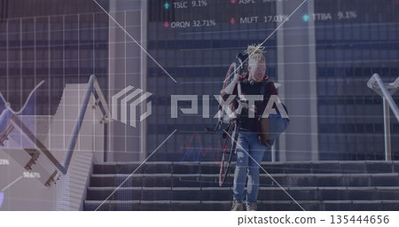 Descending cyclist carrying red and black bicycle at plaza by glass tower with financial overlays 135444656