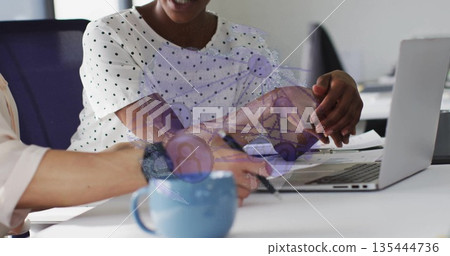 Collaborating businesswomen in suits gesturing toward laptop, taking notes at office with blue mug 135444736