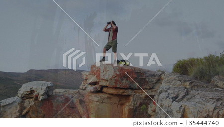 Scanning male hiker surveying canyon views from rocky cliff ledge, with binoculars and duffel bag Scanning male hiker surveying canyon views from rocky cliff ledge, with binoculars and duffel bag 135444740