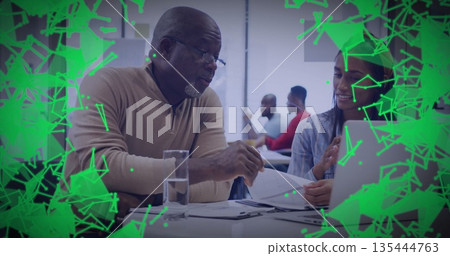 Beige-sweater mentor reviewing printed documents with mentee at office, with laptop and eyeglasses 135444763