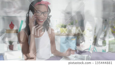 Talking Asian woman with headband reaching for laptop at office desk, with smartphone, laptop Talking Asian woman with headband reaching for laptop at office desk, with smartphone, laptop 135444881