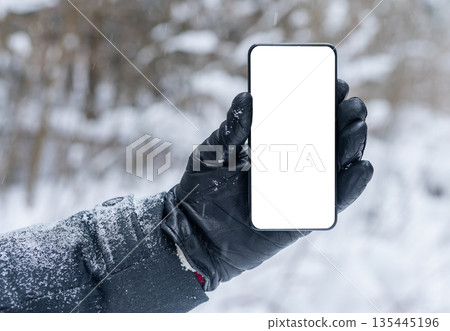 Hand in a black leather glove holding a smartphone with a blank white screen outdoors in a snowy Hand in a black leather glove holding a smartphone with a blank white screen outdoors in a snowy 135445196