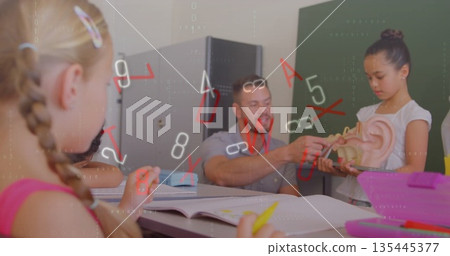 Showing mid adult male teacher pointing at anatomical ear model in classroom, with students writing 135445377