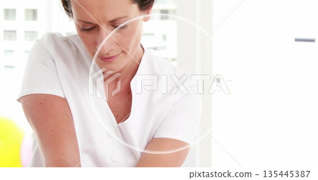 Leaning massage therapist wearing white tunic applying pressure in therapy room, with table linens 135445387