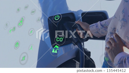 Woman in pink blouse and jeans plugging charger into car port at station, with green icons Woman in pink blouse and jeans plugging charger into car port at station, with green icons 135445412