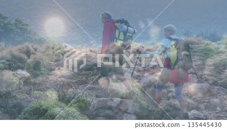 Mature couple climbing rocky hillside using trekking poles and carrying backpacks with hiking boots 135445430