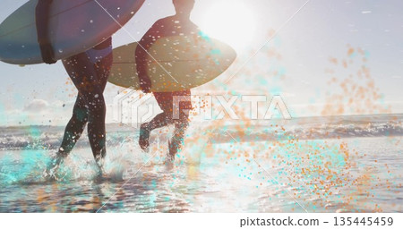 Walking surf duo carrying surfboards into ocean at beach with wetsuits, wave splashes, lens flare Walking surf duo carrying surfboards into ocean at beach with wetsuits, wave splashes, lens flare 135445459