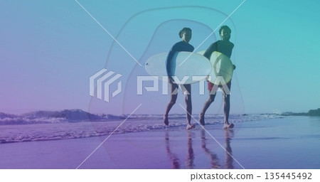 Walking surfer couple wearing wetsuit, rash guard carrying surfboards at shoreline, reflecting sky 135445492