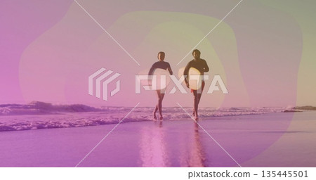 Walking couple wearing wetsuits and carrying white surfboards along sandy beach, mirrored wet sand Walking couple wearing wetsuits and carrying white surfboards along sandy beach, mirrored wet sand 135445501