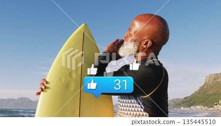 Holding yellow surfboard, senior man in wetsuit top gesturing at beach with cliffside buildings 135445510