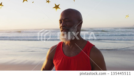 Meditating older man in red sleeveless top on beach, with golden star decorations floating overhead 135445511