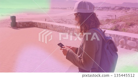 Checking smartphone woman wearing cap and backpack on promenade by stone wall, copy space 135445512