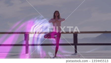 Balancing yoga practitioner performing tree pose on wooden pier by water, with digital light swirl 135445513
