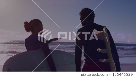 Watching surfers wearing wetsuits and shorts carrying longboard and surfboard at sunset on beach 135445529
