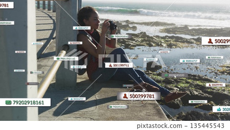 Sitting Hispanic woman holding DSLR camera and framing shoreline on seaside walkway, with data tags 135445543