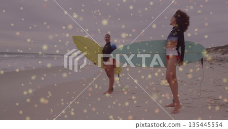 Laughing two surfers wearing gear holding yellow and pale blue surfboards at beach, with leashes 135445554
