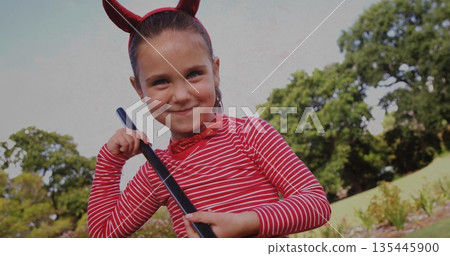 Young girl in devil costume holding broomstick, smiling playfully outdoors Young girl in devil costume holding broomstick, smiling playfully outdoors 135445900
