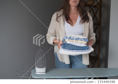 Woman irons laundry at home holding a hot iron and neatly stacking clothes showing everyday household duties concept Woman irons laundry at home holding a hot iron and neatly stacking clothes showing everyday household duties concept 135446396