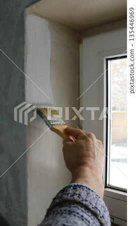 A vertical photo of a man's hand in a light sweater holding a brush and applying a layer of white paint to a window slope indoors, painting the slope of a small window in an old house 135446969