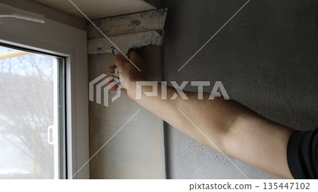 The right hand of a white man holds a wide trowel and levels the upper corner of a window opening in a room, smoothing the plaster with a spatula from the top of the window slope down The right hand of a white man holds a wide trowel and levels the upper corner of a window opening in a room, smoothing the plaster with a spatula from the top of the window slope down 135447102