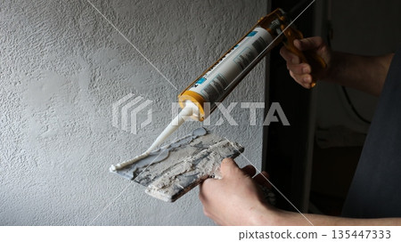 A builder's hands hold a wide spatula and a mounting gun against a gray wall, applying a bead of silicone foam to the edge of a construction knife, squeezing foam from a can onto a trowel. 135447333