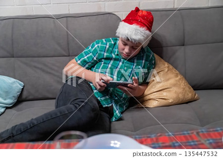 Boy in Christmas hat plays game on phone while sitting on couch at home 135447532