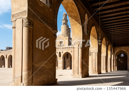 Ibn Tulun Mosque in Cairo viewed through arches. The mosque features a tall minaret and intricate architectural details. Blue sky visible in the background. 135447966