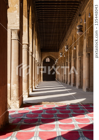 A corridor in the Ibn Tulun mosque with tall columns and intricate patterns. Soft light filters through, creating shadows. 135448043