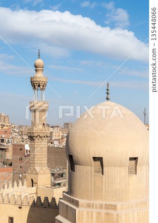 View of the Madrasa of Sarghatmish in Cairo. The scene includes domes, a minaret, and surrounding buildings under a blue sky with clouds. 135448066