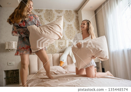 Pillow Fight Bedroom Fun: Two women playfully fight pillows on bed enjoying lighthearted fun in bedroom. Pillow Fight Bedroom Fun: Two women playfully fight pillows on bed enjoying lighthearted fun in bedroom. 135448574