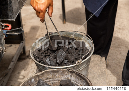 Focused man using tongs to pick up coal in workshop outdoors 135449658