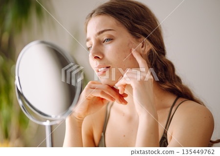 Young woman with problem skin looking into mirror. Concept of acne therapy, scars Young woman with problem skin looking into mirror. Concept of acne therapy, scars 135449762