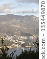 An elevated view of the popular resort town of Budva. The image captures the coastline, beaches, modern urban architecture, and majestic mountains under a cloudy sky.  135449870