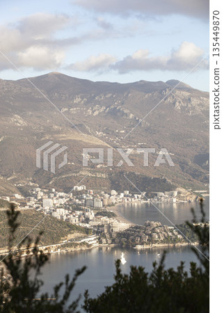 An elevated view of the popular resort town of Budva. The image captures the coastline, beaches, modern urban architecture, and majestic mountains under a cloudy sky. An elevated view of the popular resort town of Budva. The image captures the coastline, beaches, modern urban architecture, and majestic mountains under a cloudy sky. 135449870