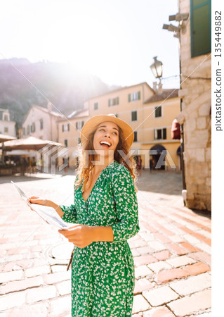 Joyful woman in a green floral dress stands in a quaint street, looking at a map. Vacation. tourism 135449882
