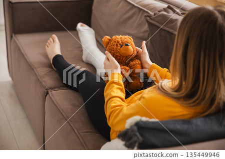 Young girl with broken leg cast sitting on sofa. Sad little kid hugging her teddy bear recovering from broken leg at home. Girl using cast and crutches. Accident injury fracture treatment foot bandage 135449946