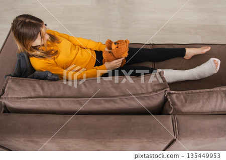 Young girl with broken leg cast sitting on sofa. Sad little kid hugging her teddy bear recovering from broken leg at home. Girl using cast and crutches. Accident injury fracture treatment foot bandage Young girl with broken leg cast sitting on sofa. Sad little kid hugging her teddy bear recovering from broken leg at home. Girl using cast and crutches. Accident injury fracture treatment foot bandage 135449953