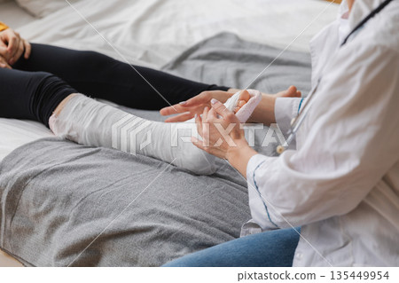 Girl with broken leg and doctor in clinic. Traumatologist orthopedist examines child leg in medical office. Doctor examining girl with medical plaster leg cast after broken ankle injury Rehabilitation 135449954