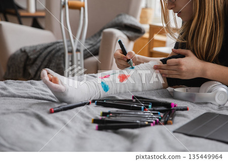 Young teenage girl with broken leg sitting on bed draws with felt tip pens on orthopedic cast. Bored girl with broken leg playing at home during rehabilitation. Accident injury fracture treatment 135449964