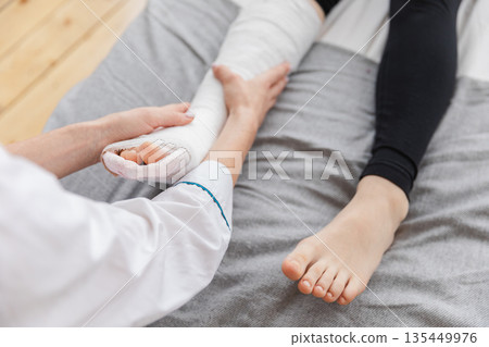 Girl with broken leg and doctor in clinic. Traumatologist orthopedist examines child leg in medical office. Doctor examining girl with medical plaster leg cast after broken ankle injury Rehabilitation 135449976