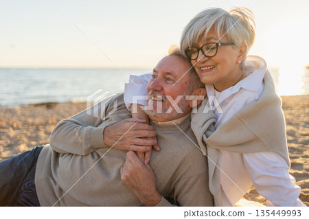 Senior mature couple sitting on beach hugging enjoying outdoor recreation. Old husband wife embracing with tenderness love enjoying sweet bonding. Grandmother grandfather together. Family moment love 135449993