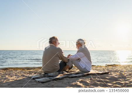Senior mature couple sitting on beach holding hands enjoying outdoor recreation. Old husband wife touching hands with tenderness love. Grandmother grandfather together. Family moment of love 135450000