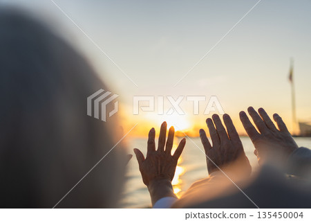 Senior couple hands at sunset. Old man woman pulls hand touching sun. Sunlight passing through fingers closeup shining sun. People stretching out hand to sun. Religion helping hand. Prayer in religion 135450004
