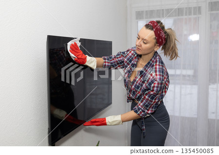 Woman Wearing Gloves Cleans a Television in Her Living Room at Daytime 135450085