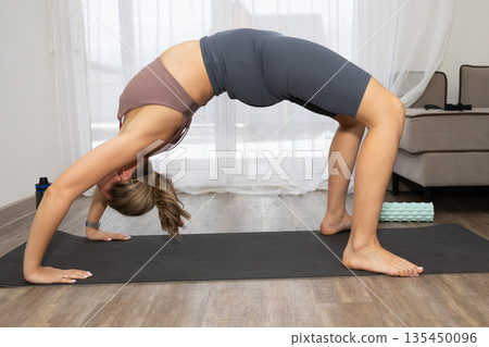 Fit Woman Performs Wheel Pose on Yoga Mat in Bright Living Room During Day 135450096