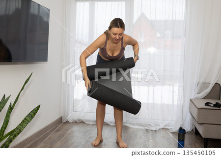 Woman Rolls up Yoga Mat in Her Living Room After Exercising Woman Rolls up Yoga Mat in Her Living Room After Exercising 135450105